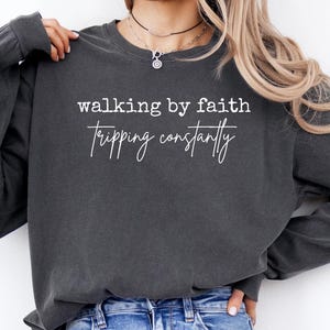 May include: Dark gray long-sleeve sweatshirt with the phrases "walking by faith" and "tripping constantly" printed in white. The sweatshirt has a crew neck and a relaxed fit, suitable for casual wear.