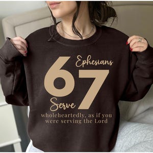 Puede incluir: Sudadera marrón oscuro con el texto "Ephesians 67 Serve" en color beige claro. El texto "wholeheartedly, as if you were serving the Lord" está impreso debajo de los números.