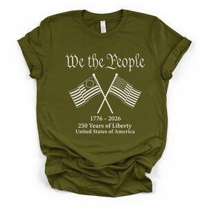 May include: Olive green t-shirt with the text "We the People" in a decorative font. Below the text are two crossed flags, one with a circle of stars and the other with stripes. The shirt also includes the text "1776-2026 250 Years of Liberty United States of America."