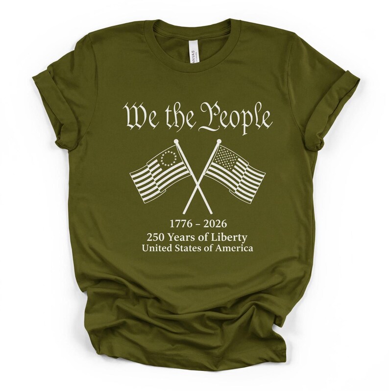 May include: Olive green t-shirt with the text "We the People" in a decorative font. Below the text are two crossed flags, one with a circle of stars and the other with stripes. The shirt also includes the text "1776-2026 250 Years of Liberty United States of America."
