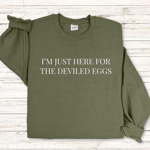 Può includere: Felpa verde oliva con la scritta bianca "I'M JUST HERE FOR THE DEVILED EGGS". La maglia a maniche lunghe ha lo scollo tondo ed è realizzata in materiale morbido.
