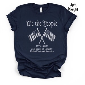 May include: Navy blue t-shirt with the text "We the People" in white script above crossed American flags. Below the flags are the dates "1776-2026" and the text "250 Years of Liberty United States of America."
