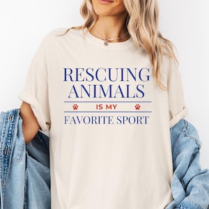 May include: Cream-colored t-shirt with the text "Rescuing Animals Is My Favorite Sport" in blue. The text is centered and includes paw print graphics. The shirt is a casual style.