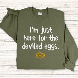 May include: Olive green sweatshirt with the text "I'm just here for the deviled eggs" in white, along with a yellow and orange deviled egg graphic. The long-sleeved shirt is folded on a light wood-grain surface.
