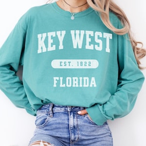 May include: Teal long-sleeve sweatshirt with the text "KEY WEST" in white block letters. Below the text is "EST. 1822" and "FLORIDA" in white block letters. The sweatshirt is made of a soft material.
