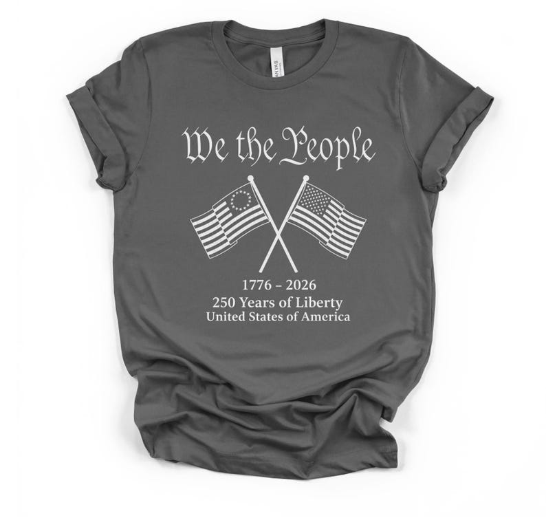 May include: Dark gray t-shirt with the text "We the People" above crossed American flags. The flags are white with blue and red accents. Below the flags is the text "1776-2026 250 Years of Liberty United States of America."