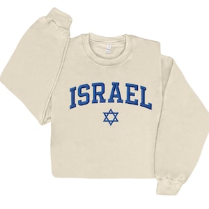 May include: Cream-colored sweatshirt with the word "ISRAEL" embroidered in blue, arched above a blue Star of David. The long-sleeved top is made of a soft material.