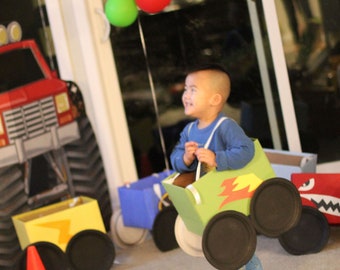 May include: A young child dressed in a blue shirt is playing with a cardboard monster truck toy. The truck is green, yellow, and black with a lightning bolt design. The child is holding the steering wheel and looking to the right. There are balloons in the background.