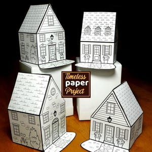 以下が含まれることがあります： 詳細な建築デザインが施された、白黒の紙製の家のモデルが4つ。各家には窓、ドア、屋根のディテールが施されています。家は木製の表面に展示されており、茶色のボックスに「Timeless paper Project」というテキストが表示されています。