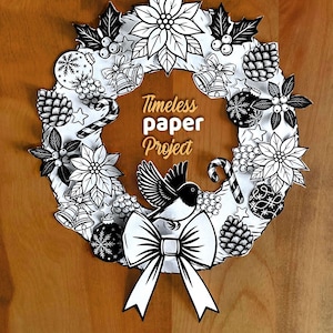 May include: A black and white paper wreath featuring illustrations of poinsettias, pinecones, bells, candy canes, and ornaments. A bird is centred at the bottom above a large bow. The text Timeless paper Project is printed in the centre.