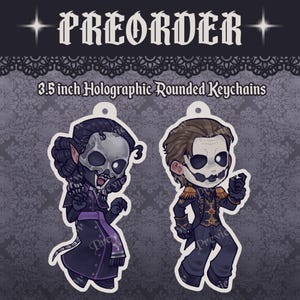 May include: Two 3.5 inch holographic rounded keychains featuring stylized characters. The left character wears a dark robe with a skull mask, while the right character wears a dark military-style jacket with gold epaulets and a skull mask.