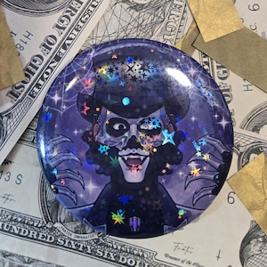 May include: A round, holographic button featuring a stylized illustration of a skeletal figure with a top hat and clawed hands. The design incorporates stars and spiderwebs, with a purple and black color scheme. The button is set against a backdrop of dollar bills.