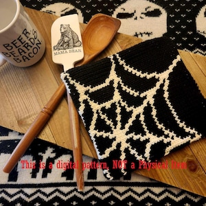 May include: A black and white crochet pot holder with a spiderweb design. The pot holder is on a wooden surface with a wooden spoon and a spatula with a bear design. The text "BEER BEARDS BACON" is on a white mug in the background. The text "MAMA BEAR" is on the spatula.