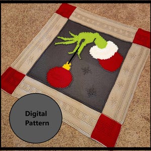 Puede incluir: Una manta de crochet con una mano verde del Grinch sosteniendo un adorno navideño rojo y blanco. La manta tiene un centro gris con un borde beige y esquinas rojas. El texto "Patrón digital" está escrito en un círculo en la esquina inferior izquierda de la manta.