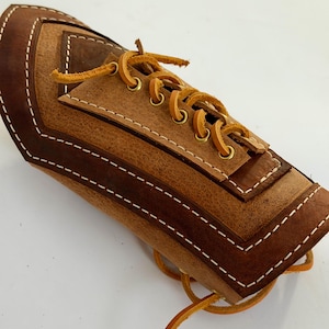 May include: A brown leather bracer with a laced closure. The bracer features a layered design with darker brown accents and white stitching. The laces are a lighter brown color.