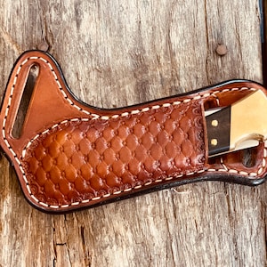May include: A brown leather knife sheath with a diamond-patterned centre and white stitching. A partially visible knife with a gold blade and black handle is in the sheath. The sheath has a belt loop.