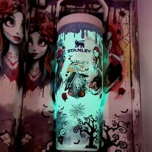 Puede incluir: Un termo Stanley con un dise&ntilde;o que brilla en la oscuridad con personajes de "Pesadilla antes de Navidad". El dise&ntilde;o incluye a Jack Skellington, Sally y un &aacute;rbol espeluznante. El termo tiene un asa blanca y un brillo azul claro.