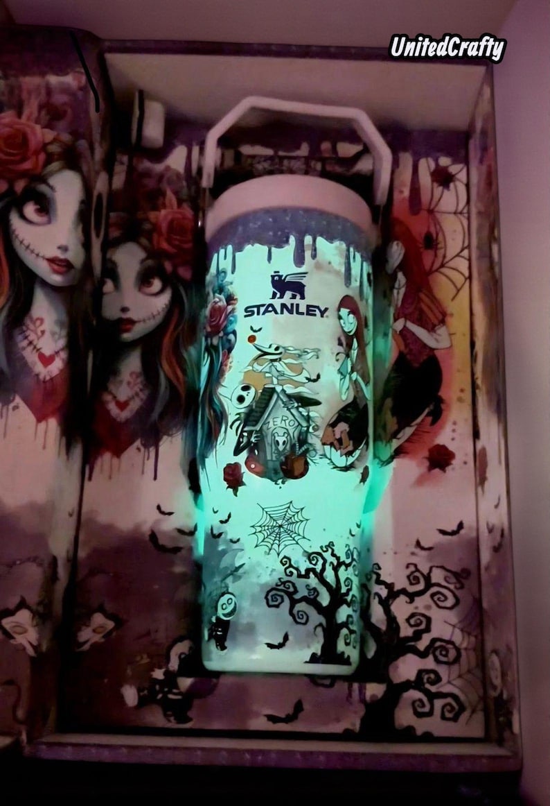 Puede incluir: Un termo Stanley con un dise&ntilde;o que brilla en la oscuridad con personajes de "Pesadilla antes de Navidad". El dise&ntilde;o incluye a Jack Skellington, Sally y un &aacute;rbol espeluznante. El termo tiene un asa blanca y un brillo azul claro.
