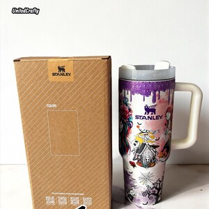 Puede incluir: Un vaso Stanley con asa y tapa blancas, con un dise&ntilde;o colorido con motivos de Halloween. El vaso est&aacute; junto a una caja marr&oacute;n lisa con el logotipo de Stanley.
