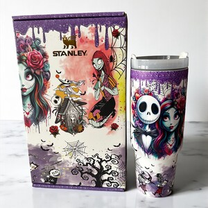 Puede incluir: Un vaso Stanley y su caja a juego presentan un dise&ntilde;o espeluznante y colorido. El vaso y la caja muestran ilustraciones de personajes de Pesadilla antes de Navidad, con un esquema de color p&uacute;rpura y blanco.