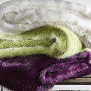 May include: Three faux fur throws stacked on top of each other. The top throw is white, the middle throw is green, and the bottom throw is purple.