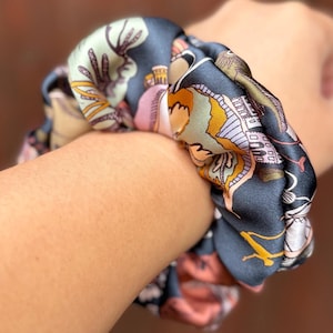 Könnte beinhalten: Ein dunkelblaues Scrunchie mit einem farbenfrohen Muster aus Gebäuden, Blumen und anderen Designs. Das Scrunchie ist um ein Handgelenk gewickelt und zeigt die Details und Farben des Stoffes. Der Hintergrund ist ein verschwommenes Rotbraun.