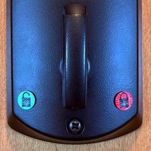 May include: A black door handle with a green and red sticker on it. The stickers have a padlock icon on them.