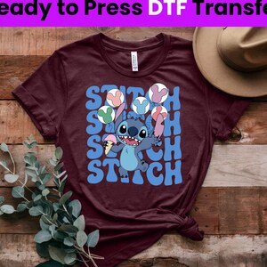 Puede incluir: Una camiseta color granate con un gr&aacute;fico azul y blanco de Stitch de Lilo & Stitch. El gr&aacute;fico muestra a Stitch sosteniendo un cono de helado y rodeado de globos de colores. El texto "Stitch" se repite en una fuente retro.