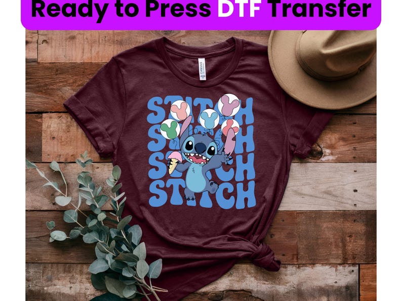 Puede incluir: Una camiseta color granate con un gr&aacute;fico azul y blanco de Stitch de Lilo & Stitch. El gr&aacute;fico muestra a Stitch sosteniendo un cono de helado y rodeado de globos de colores. El texto "Stitch" se repite en una fuente retro.