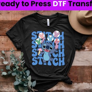 Puede incluir: Camiseta negra con un personaje azul Stitch sosteniendo un cono de helado y rodeado de globos de colores. El texto "Stitch" est&aacute; escrito en letras azules en la camiseta.