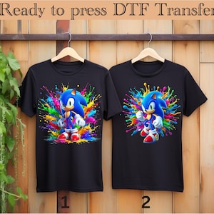 May include: Two black t-shirts with a colorful paint splatter design featuring Sonic the Hedgehog. The shirts are labeled 1 and 2.