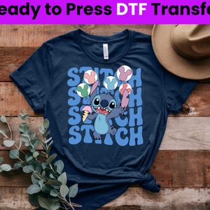 Puede incluir: Una camiseta azul marino con un gr&aacute;fico de Stitch de Lilo & Stitch sosteniendo un helado y haciendo malabares con orejas de Mickey Mouse. El texto "Stitch" se repite en una fuente retro.