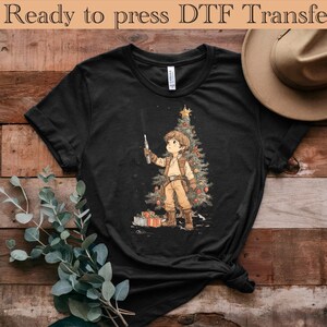 以下が含まれることがあります： 黒いTシャツ。クリスマスツリーの前でライトセーバーを持っているキャラクターの漫画イラスト。キャラクターはベージュの衣装を着ています。「Ready to press DTF Transfer」の文字が上部にあります。