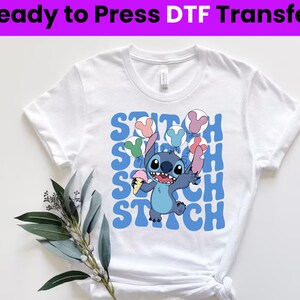 Puede incluir: Camiseta blanca con un gr&aacute;fico azul y blanco de Stitch de Lilo & Stitch sosteniendo un cono de helado azul y globos. El texto "Stitch" se repite en una fuente retro azul.