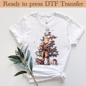 以下が含まれることがあります： クリスマスのテーマのイラストが描かれた白いTシャツ。イラストには、飾られたクリスマスツリーの前に立っている漫画のキャラクターが描かれています。キャラクターはプレゼントボックスを持っており、赤と白のストライプの帽子をかぶっています。