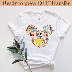 以下が含まれることがあります： ミッキーマウスの頭の形に配置されたディズニーキャラクターのカラフルなグラフィックが特徴の白いTシャツ。デザインには白雪姫、七人の小人、その他のキャラクターが含まれています。「Ready to press DTF Transfer」というテキストが上部にあります。