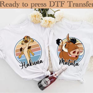 May include: Two white t-shirts featuring a graphic print of characters from the film The Lion King. The shirt on the left has a graphic of Timon the meerkat with the text "Hakuna Matata". The shirt on the right has a graphic of Pumbaa the warthog with the text "Hakuna Matata".