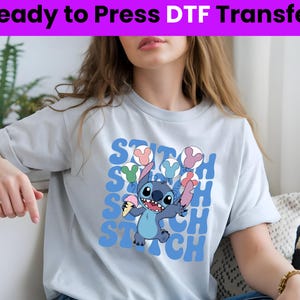 Puede incluir: Una camiseta azul claro con un estampado gr&aacute;fico de Stitch de Lilo & Stitch sosteniendo un cono de helado. El gr&aacute;fico est&aacute; rodeado por la palabra "Stitch" repetida en una fuente retro colorida.