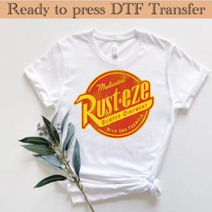 Puede incluir: Camiseta blanca con un gráfico rojo y amarillo de un producto de automóvil vintage llamado "Rust-eze Medicated Bumper Ointment Rear End Formula".