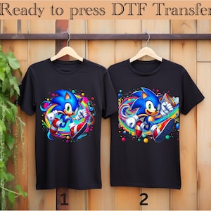 May include: Two black t-shirts with a colorful graphic of Sonic the Hedgehog running. The graphic includes a rainbow and colorful paint splatters. The t-shirts are labeled 1 and 2.