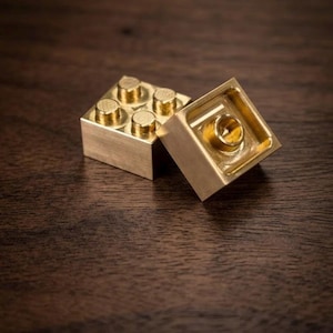 May include: Two metallic, gold-colored building blocks. One block is positioned upright, displaying four circular studs on its top surface. The second block is tilted, revealing its hollow underside with a single central circular connector.