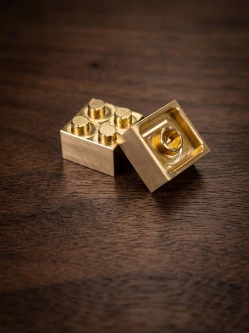 May include: Two metallic, gold-colored building blocks. One block is positioned upright, displaying four circular studs on its top surface. The second block is tilted, revealing its hollow underside with a single central circular connector.