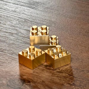 May include: Three metallic gold-colored building blocks, each featuring a square base and four cylindrical studs on the top surface. The blocks are arranged in a cluster, showcasing a smooth, reflective finish and a solid, blocky construction.