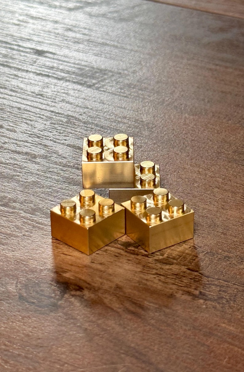 May include: Three metallic gold-colored building blocks, each featuring a square base and four cylindrical studs on the top surface. The blocks are arranged in a cluster, showcasing a smooth, reflective finish and a solid, blocky construction.