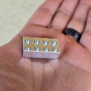 May include: A small, shiny, silver-colored building block with eight circular studs on top. The rectangular block is held in an open hand, showcasing its metallic finish and precise construction. A classic toy reimagined.