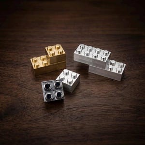 May include: A collection of four interlocking building blocks made of solid metal. One block is finished in a warm brass tone, while the other three are finished in a cool silver tone. Each block features a standard rectangular shape with circular studs.