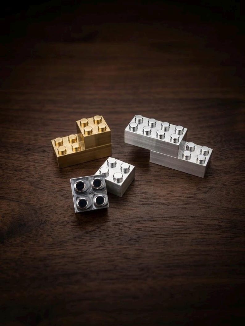 May include: A collection of four interlocking building blocks made of solid metal. One block is finished in a warm brass tone, while the other three are finished in a cool silver tone. Each block features a standard rectangular shape with circular studs.