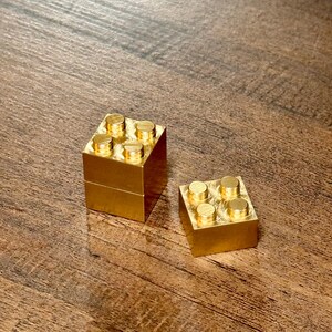 May include: Two metallic gold-colored building blocks, each featuring a square base and four circular studs on the top surface. One block is stacked vertically on top of another, while the second block sits flat beside it on the surface.