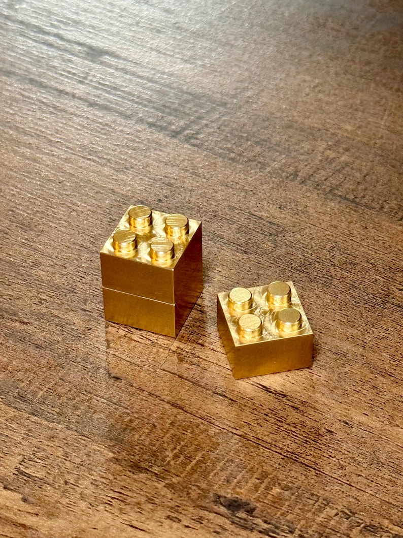 May include: Two metallic gold-colored building blocks, each featuring a square base and four circular studs on the top surface. One block is stacked vertically on top of another, while the second block sits flat beside it on the surface.