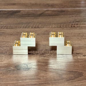 May include: Two metallic gold-colored building blocks, each featuring a rectangular base with four circular studs on the top surface. The blocks are arranged in a stepped configuration, showcasing a smooth, reflective finish and precise geometric edges.